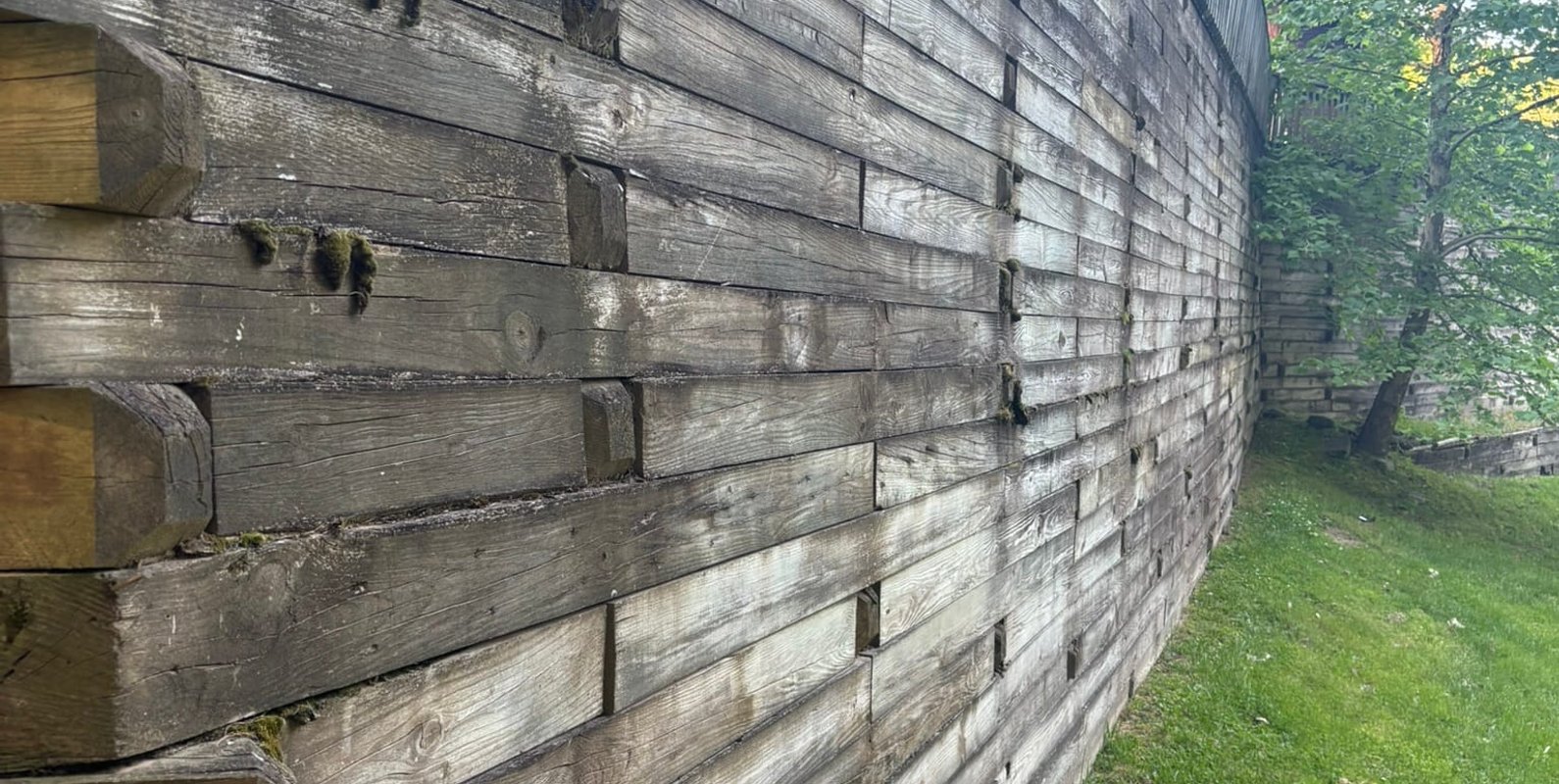 A tall wood retaining wall showing surface weathering, minor joint gaps, and early signs of outward movement—conditions that indicate aging materials and increasing lateral pressure behind the wall.